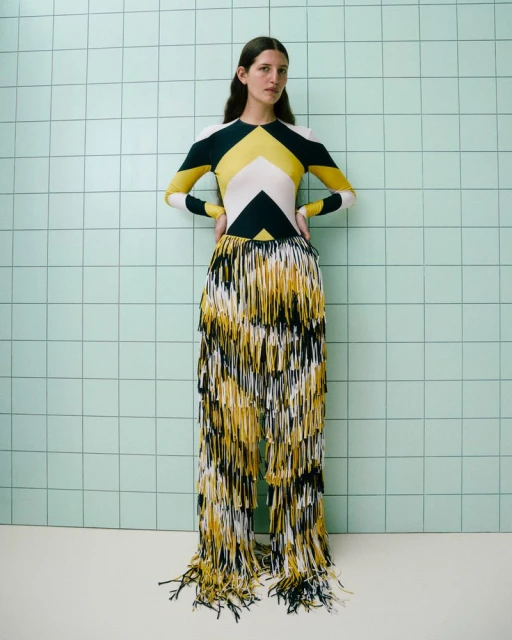 A woman poses indoors wearing a yellow, white, and black geometric pattern top, and yellow, white, and black fringe pants.