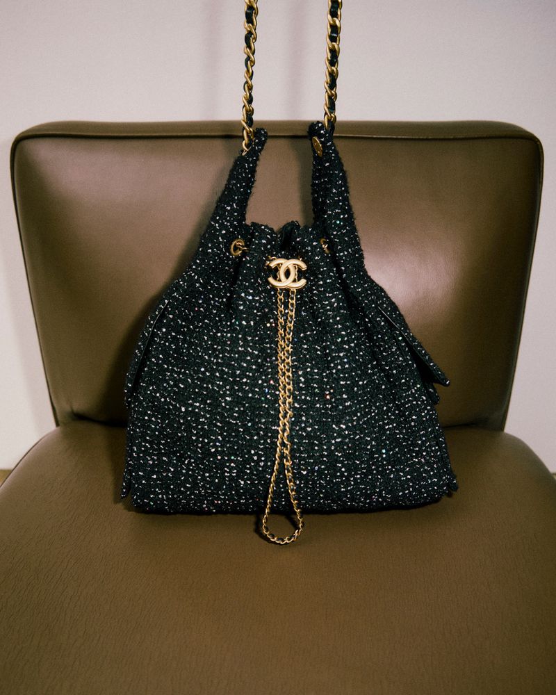 A black tweed Chanel handbag with a gold chain strap sits on a brown leather chair in a studio setting with neutral lighting.