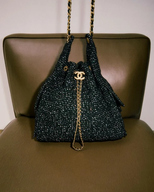 A black tweed Chanel handbag with a gold chain strap sits on a brown leather chair in a studio setting with neutral lighting.