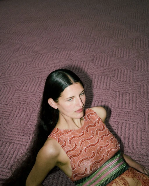 Fashion shot of a woman in an orange dress lying on a textured mauve surface, captured in a medium shot with soft, diffused lighting.