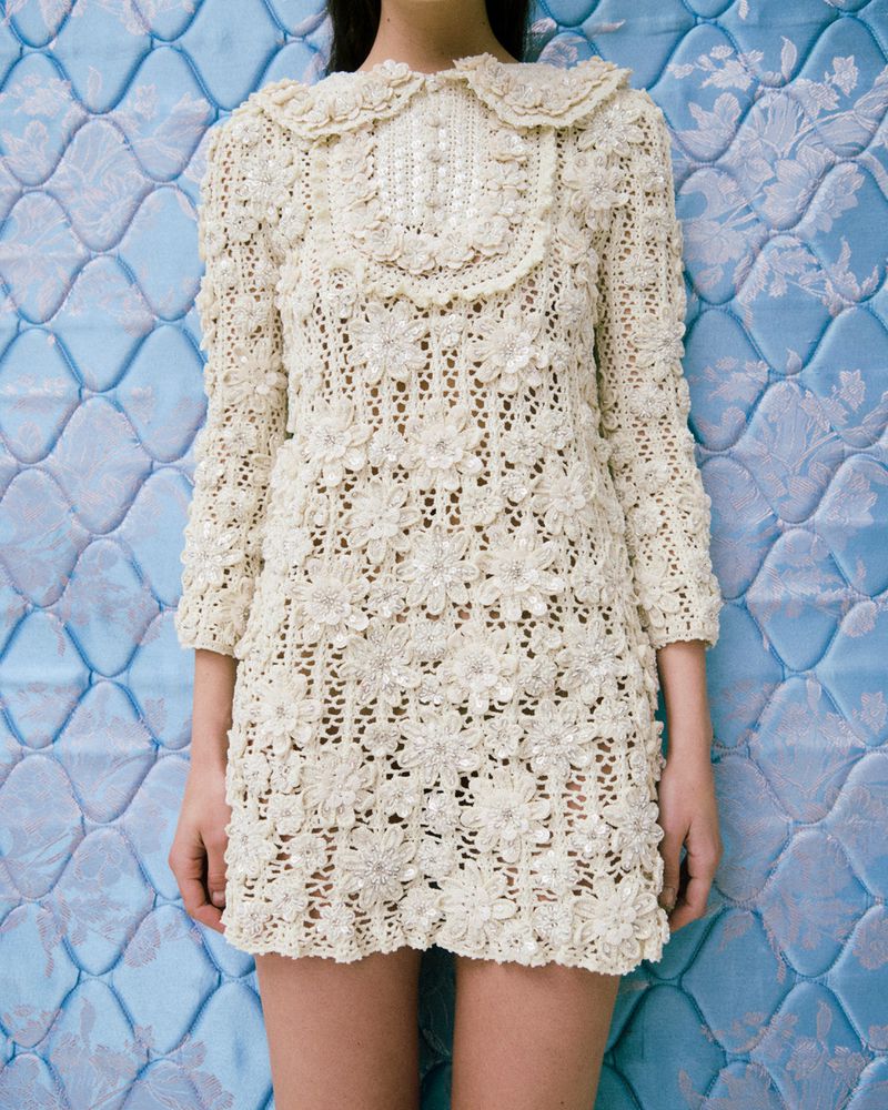 Close-up of a slender model wearing a short, cream-colored, floral-patterned lace dress, posing against a textured blue background.