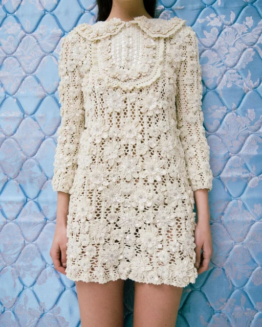 Close-up of a slender model wearing a short, cream-colored, floral-patterned lace dress, posing against a textured blue background.