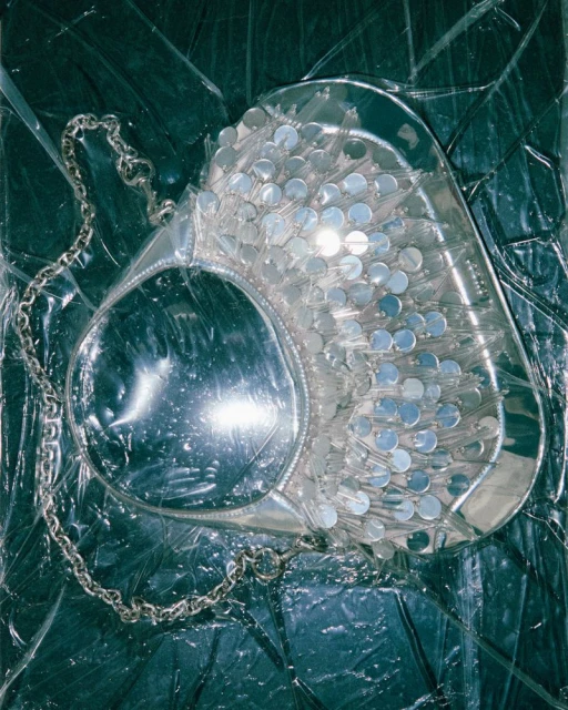 An overhead shot of a shiny, transparent handbag with a chain handle, set against a background of crumpled, translucent plastic.