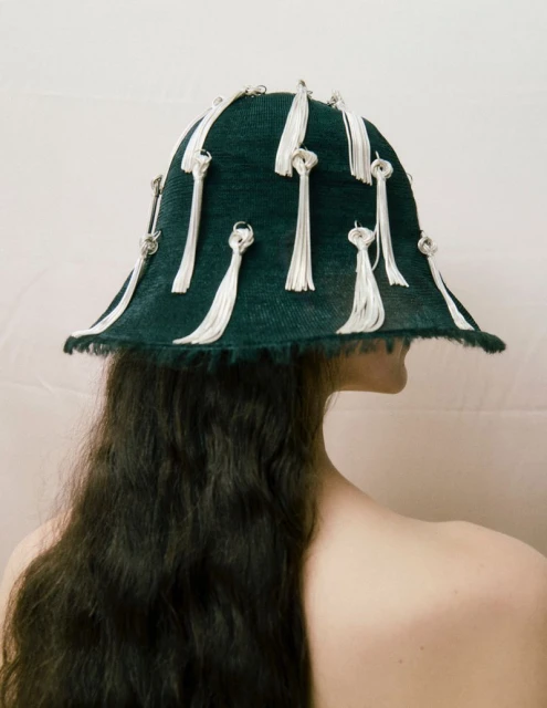 Back view of a person with long dark hair wearing a green hat with white tassels against a white background in a studio setting.