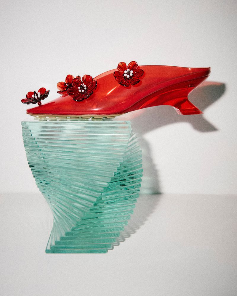 Elevated view of a red transparent shoe decorated with flowers displayed on a clear acrylic pedestal against a white background.