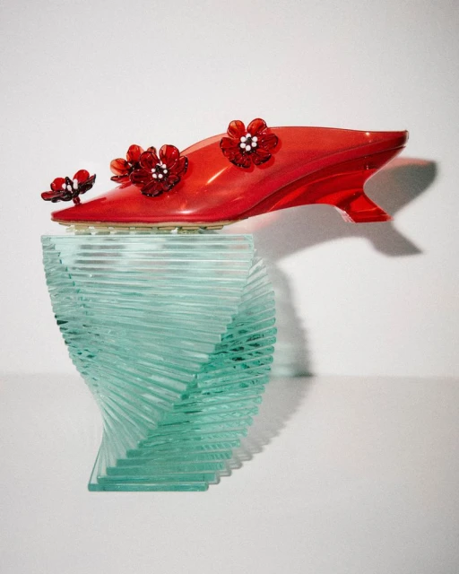 Elevated view of a red transparent shoe decorated with flowers displayed on a clear acrylic pedestal against a white background.