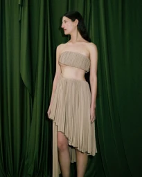 Model wearing a pleated beige dress standing against dark green draped fabric background