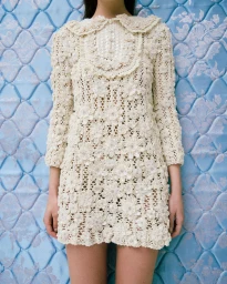 Model wearing a heavily textured cream-colored lace dress standing against a blue quilted background.