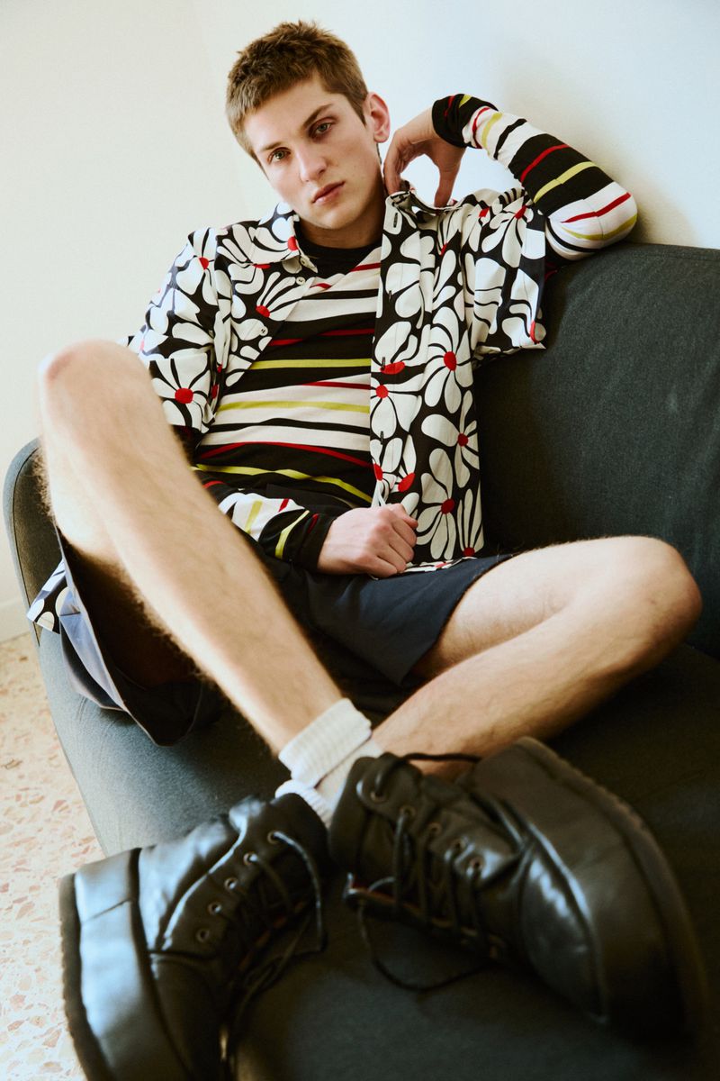 A young man with light brown hair poses on a dark couch, wearing a black and white flower shirt, black shorts, and chunky platform shoes.