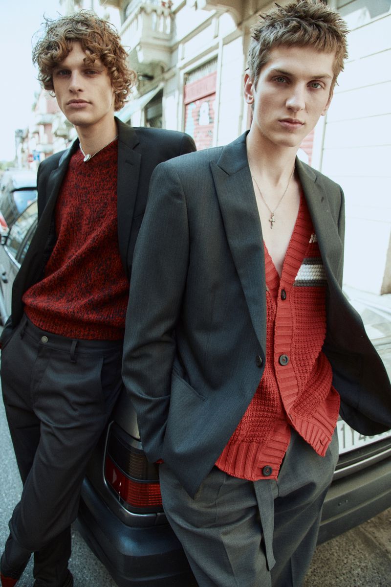 Two young adult men pose in a city street, dressed in stylish blazers and sweaters, leaning against a car with urban buildings visible.