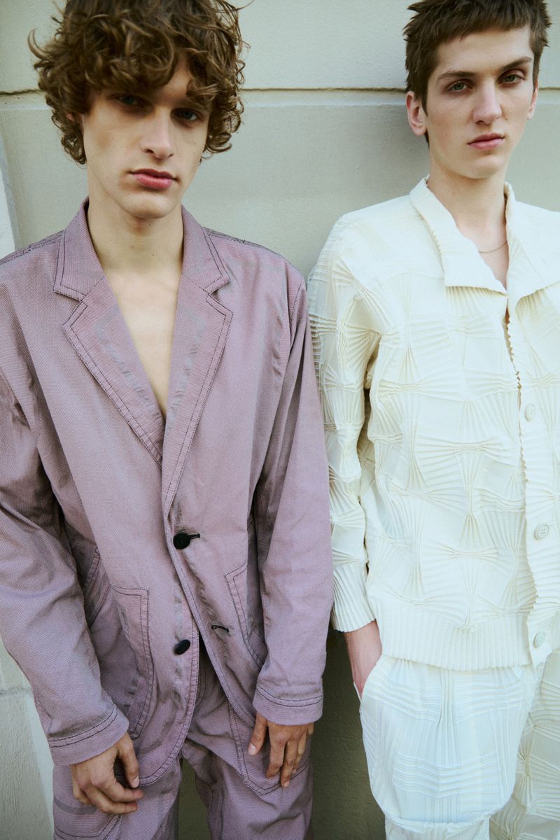 Two men model fashion apparel outdoors. One wears a mauve suit, and the other a cream textured set. Neutral background.