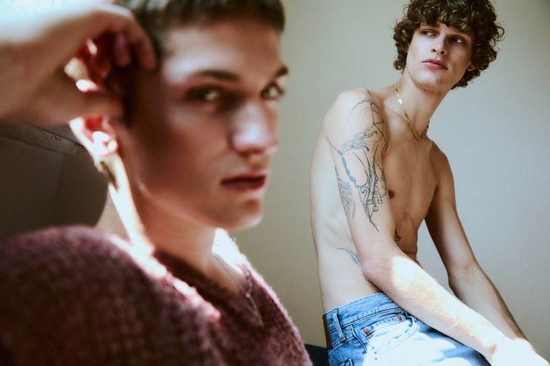 Two light-skinned adult males pose indoors in a natural light setting. One wears a sweater and the other is shirtless with jeans and tattoos.
