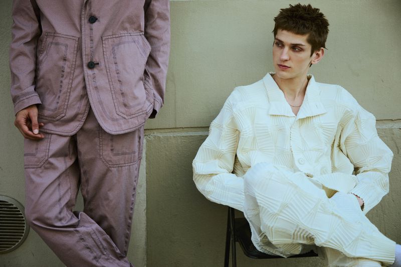 Two fashion models in matching tailored outfits pose against a neutral wall, highlighting textural details and modern tailoring.