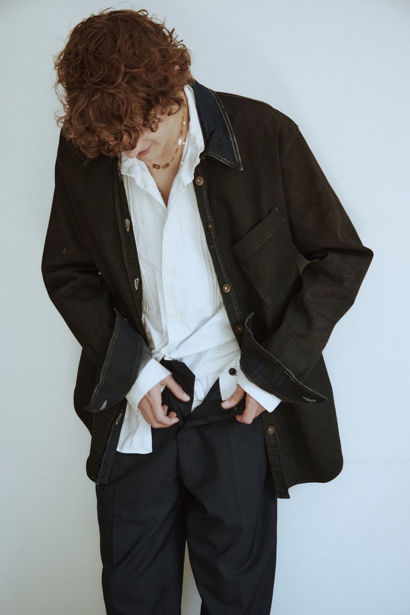 Adult male with curly hair wearing a black jacket and white shirt against a white background, adjusting his black pants.