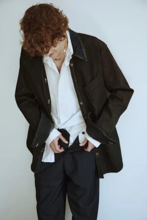 Adult male with curly hair wearing a black jacket and white shirt against a white background, adjusting his black pants.