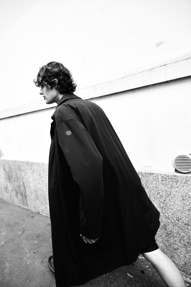 A young adult male with curly hair wearing a long black coat walks along a city street next to a white wall on an overcast day.
