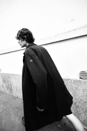 Monochrome fashion editorial image of a male model walking outdoors in a long dark coat alongside a stone building.