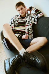 Young male model seated on a dark sofa wearing a patterned floral shirt, striped t-shirt, shorts, and chunky black boots.