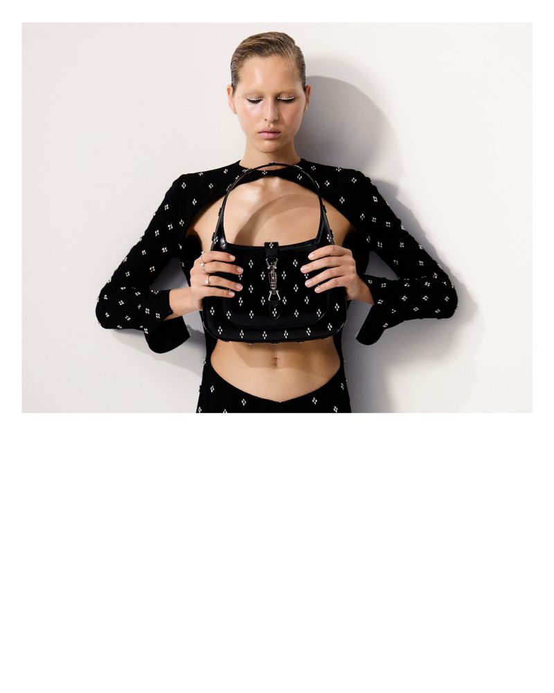 A slender woman with a sleek hairstyle poses against a white wall, holding a black, embellished purse. She's wearing matching cutout clothing.