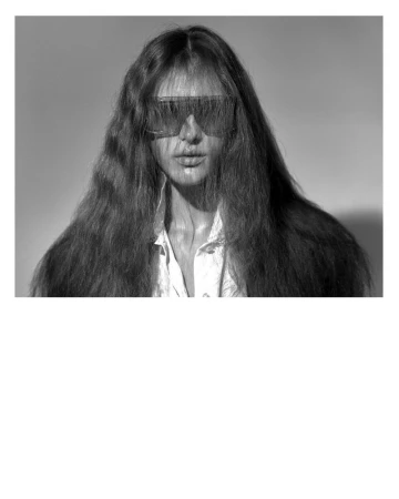 Model Angelina Kendall in sunglasses with long dark hair and a white collared shirt against a grey background