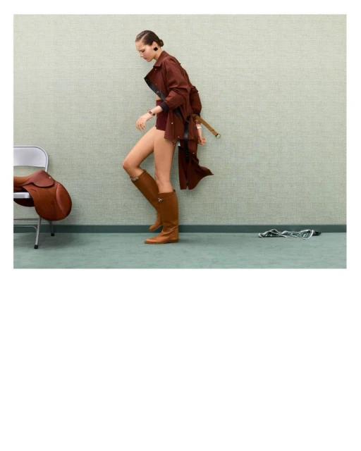 A woman in a brown coat and boots walks past a chair with a saddle in an indoor setting with a textured light green wall and flooring.