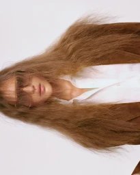 Close-up portrait of a model with long brown hair wearing tinted glasses, a white button-down shirt, and a brown jacket against a white background.