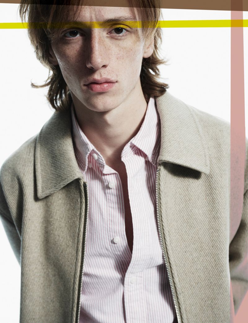 Close-up of a young adult male with light brown hair wearing an open pink and white striped shirt under a zip-up beige jacket in a studio.
