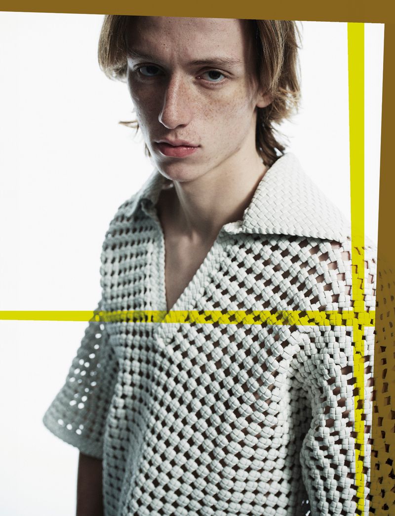 Studio shot of a young adult male wearing a white crochet shirt, against a white background with abstract gold accents.