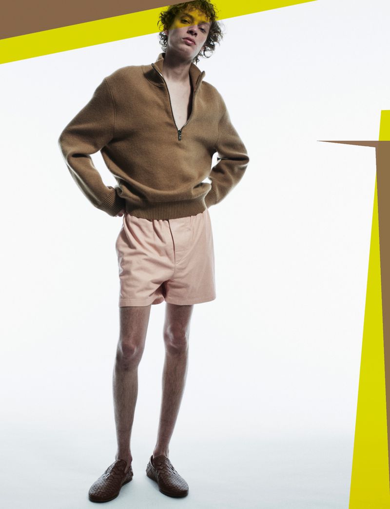 Fashion shot of a slender adult male model posing in a brown knit quarter-zip sweater and pink shorts against a white background with yellow accents.
