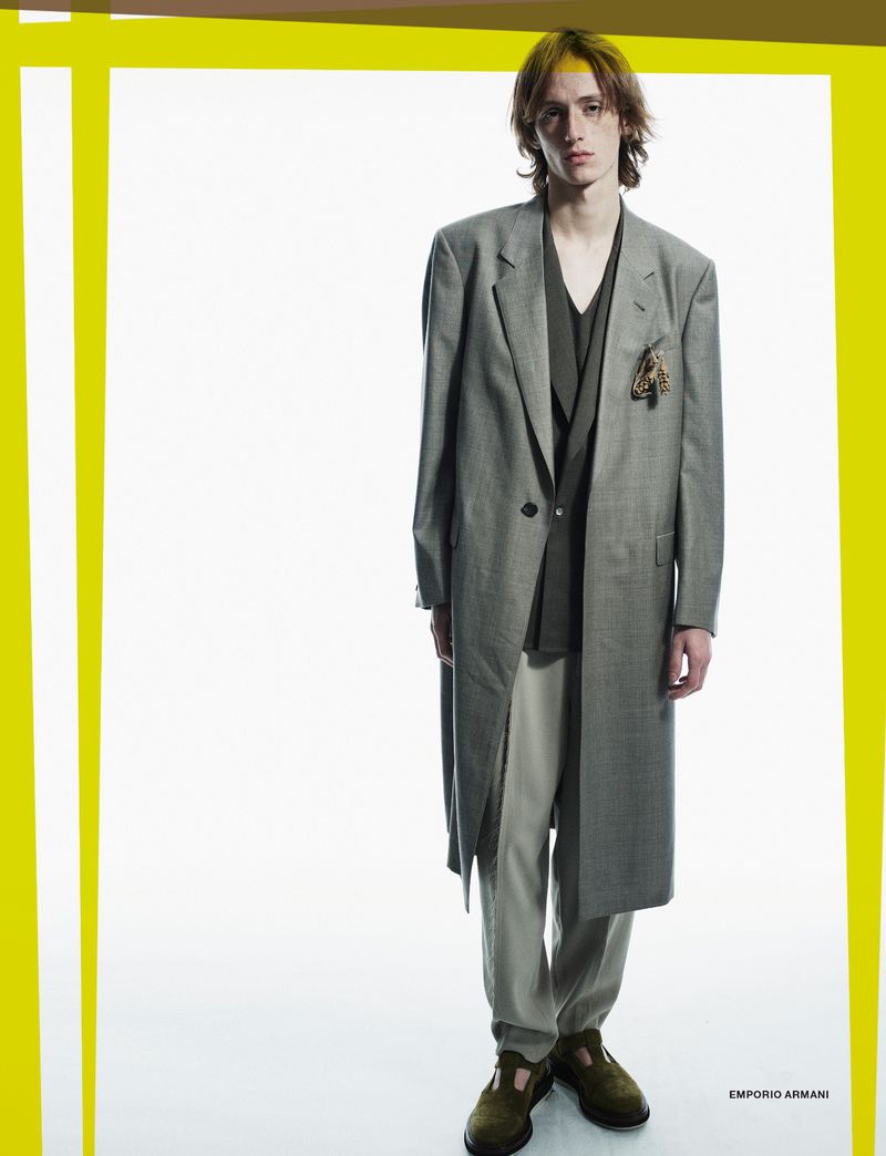 A young adult male model poses in a full-length shot, wearing a grey overcoat, a dark-green shirt, beige pants, and dark-green shoes on a white backdrop.