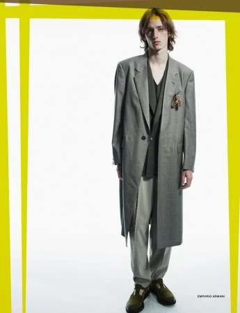 A young adult male model poses in a full-length shot, wearing a grey overcoat, a dark-green shirt, beige pants, and dark-green shoes on a white backdrop.