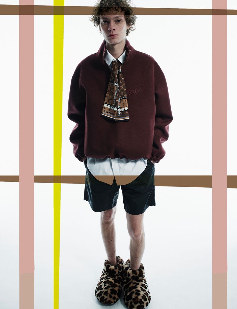 Full-length shot of a young adult man in a burgundy sweatshirt, tie, shorts, and leopard print slippers, posed against a white backdrop.
