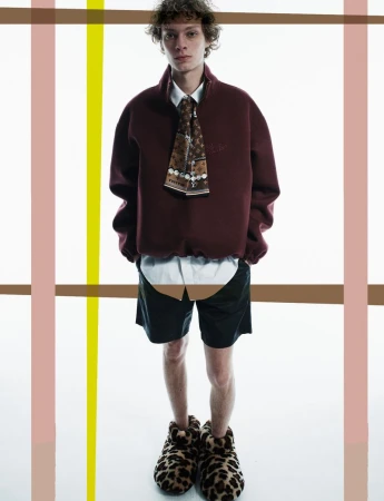 Full-length shot of a young adult man in a burgundy sweatshirt, tie, shorts, and leopard print slippers, posed against a white backdrop.