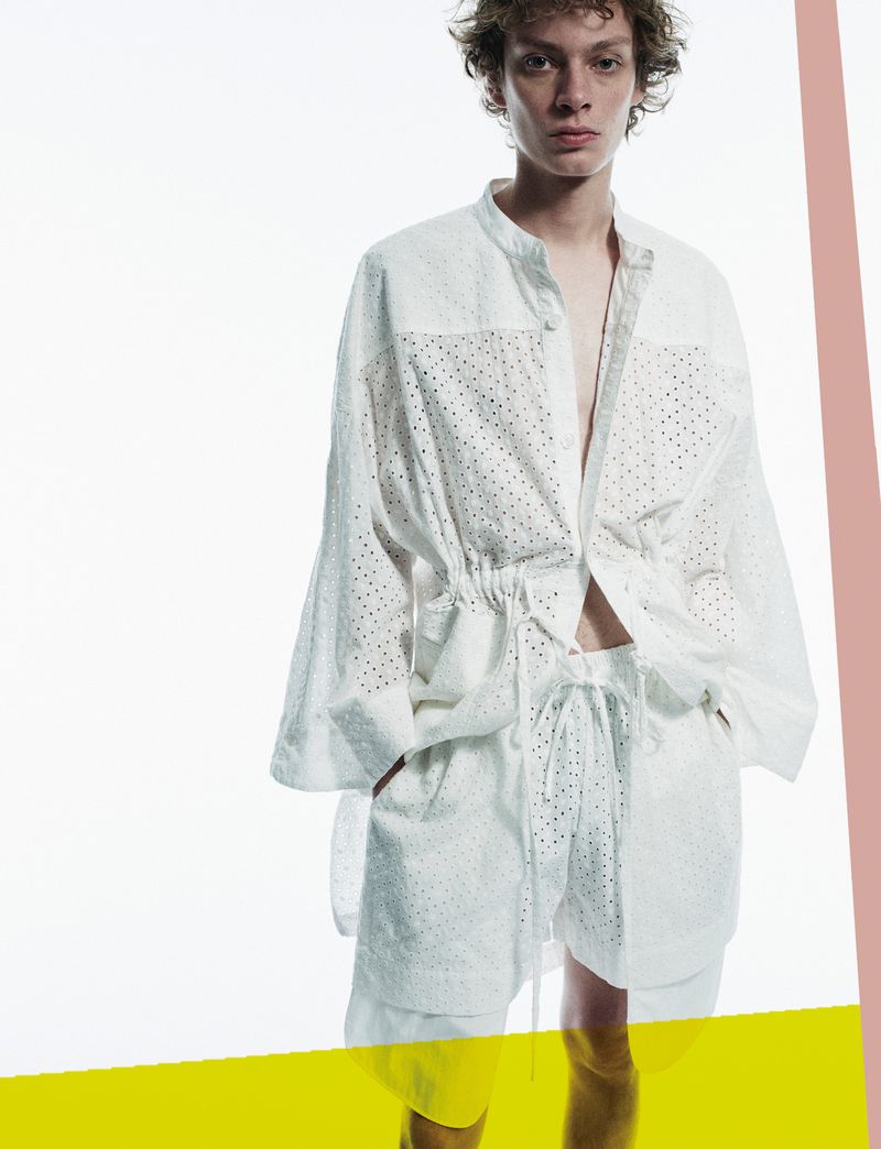 Young adult male model posing in a white, semi-sheer, long-sleeved shirt and matching shorts in a bright studio environment.