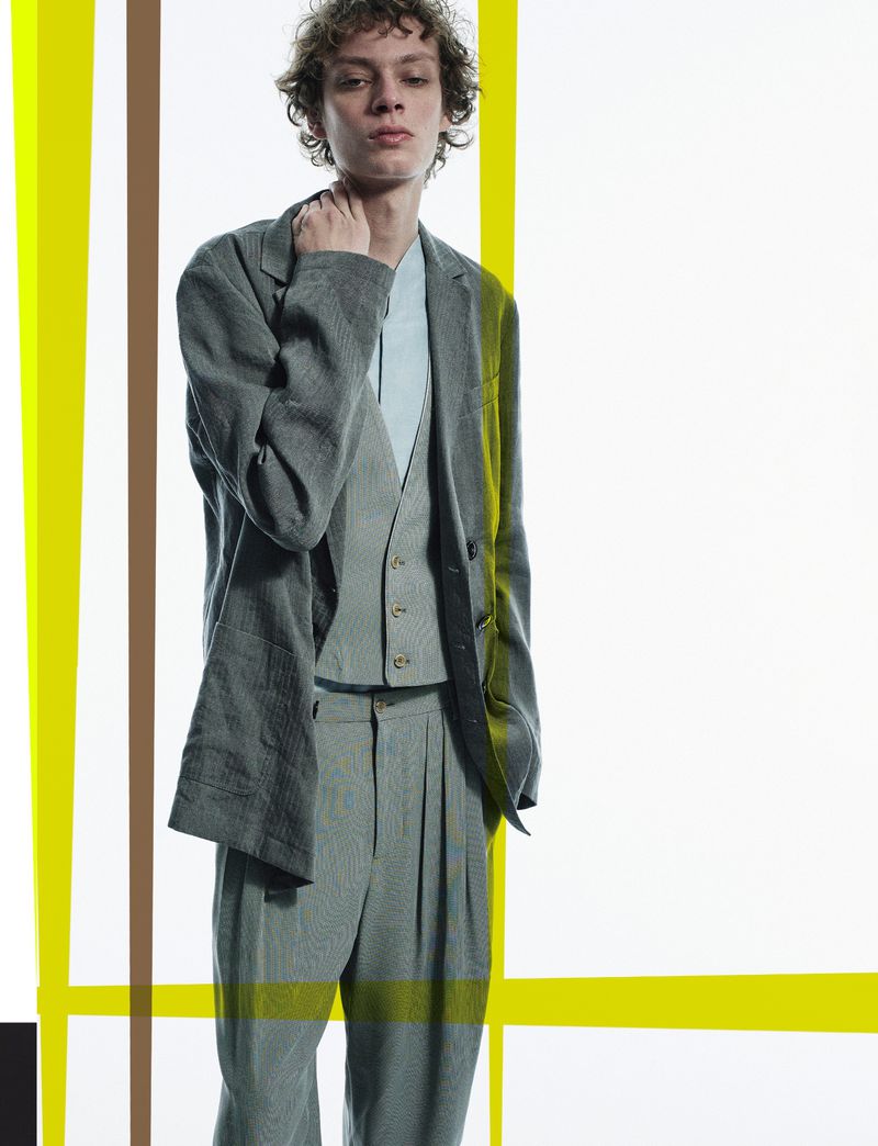 Portrait of an adult male in a grey tailored suit, vest, and blue shirt against a studio backdrop with yellow and brown vertical lines.