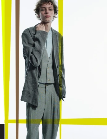 Portrait of an adult male in a grey tailored suit, vest, and blue shirt against a studio backdrop with yellow and brown vertical lines.