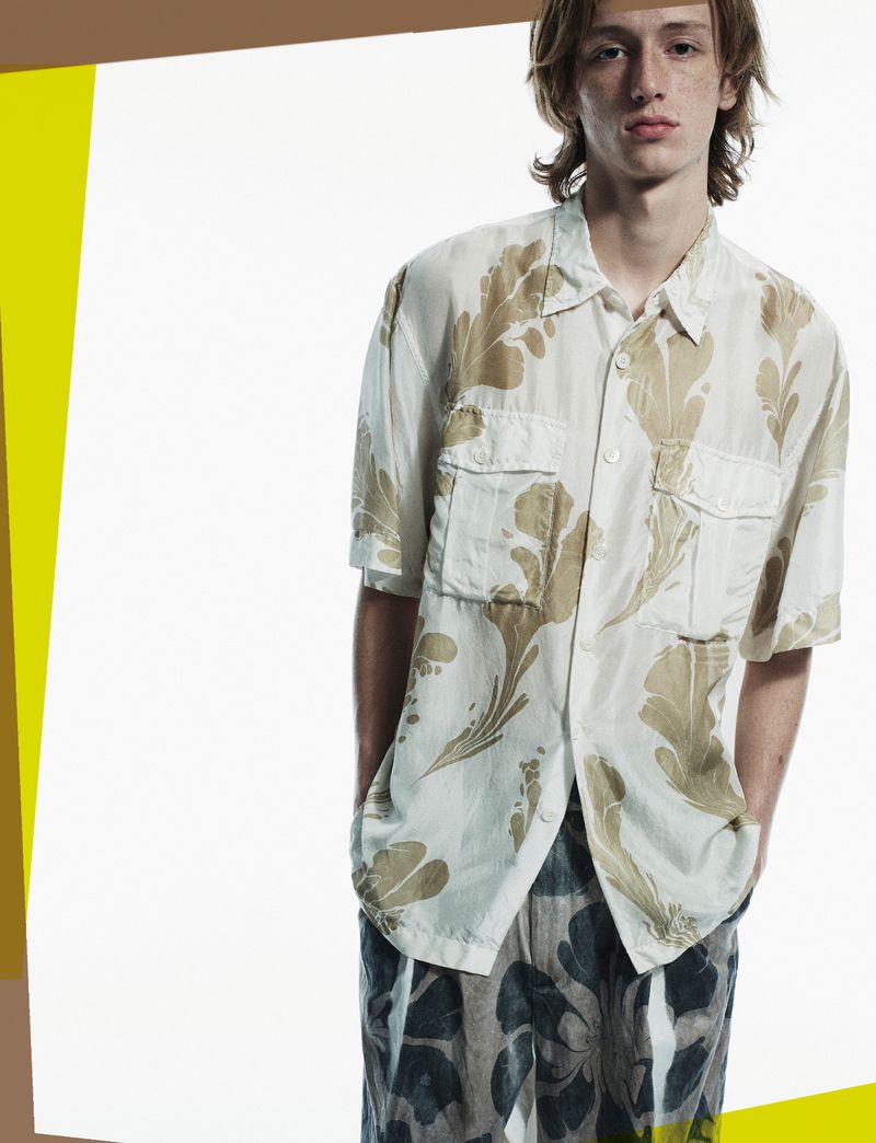 Young adult male in a beige floral-patterned shirt and floral pants, standing indoors in a full shot against a neutral background.