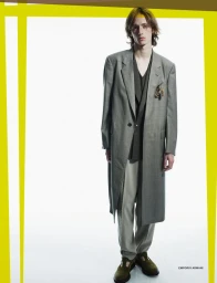 Model standing against a white seamless background wearing a grey overcoat and trousers.