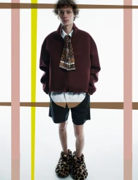 Young male model wearing burgundy fleece, white shirt, patterned scarf and leopard print slippers against a plain background