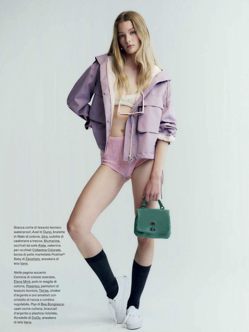 Fashion editorial shot of a fair-skinned woman in a lilac jacket and pink shorts, accessorized with a small green handbag and black socks.