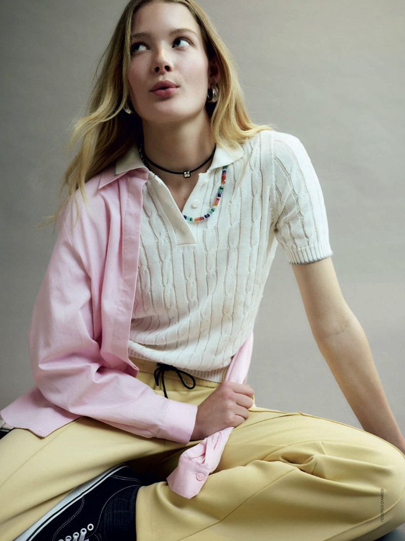 Blonde-haired woman in a white knit polo and yellow pants, posing indoors against a light gray backdrop. She wears a colorful necklace.
