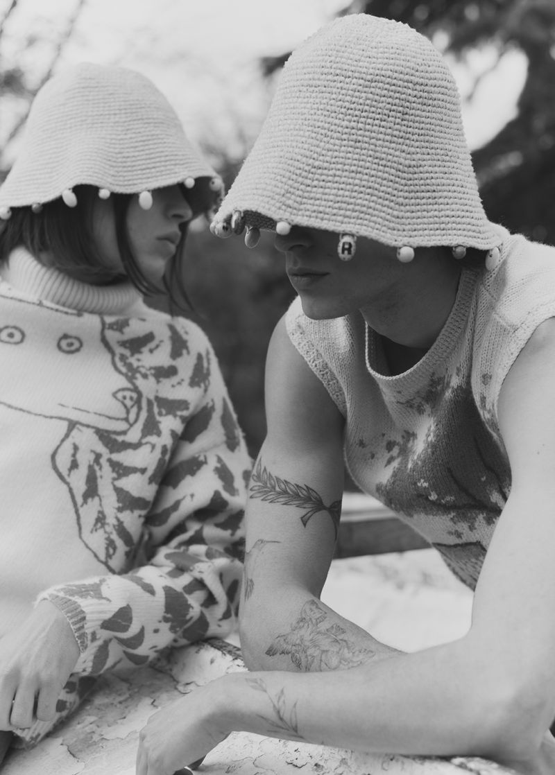 Two people wearing hats with beaded trim, one in patterned sweater, outdoor setting in black and white, showcasing textures and unique style.