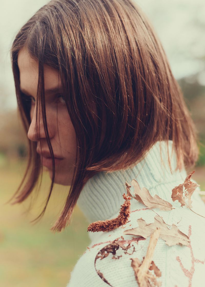 Close-up portrait of a male model with shoulder-length brown hair wearing a light blue turtleneck sweater adorned with fallen autumn leaves.