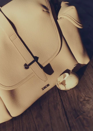 Beige leather backpack and damaged eggshell on a dark wood surface. Flat lighting emphasizes textures and shadows in a still life composition.
