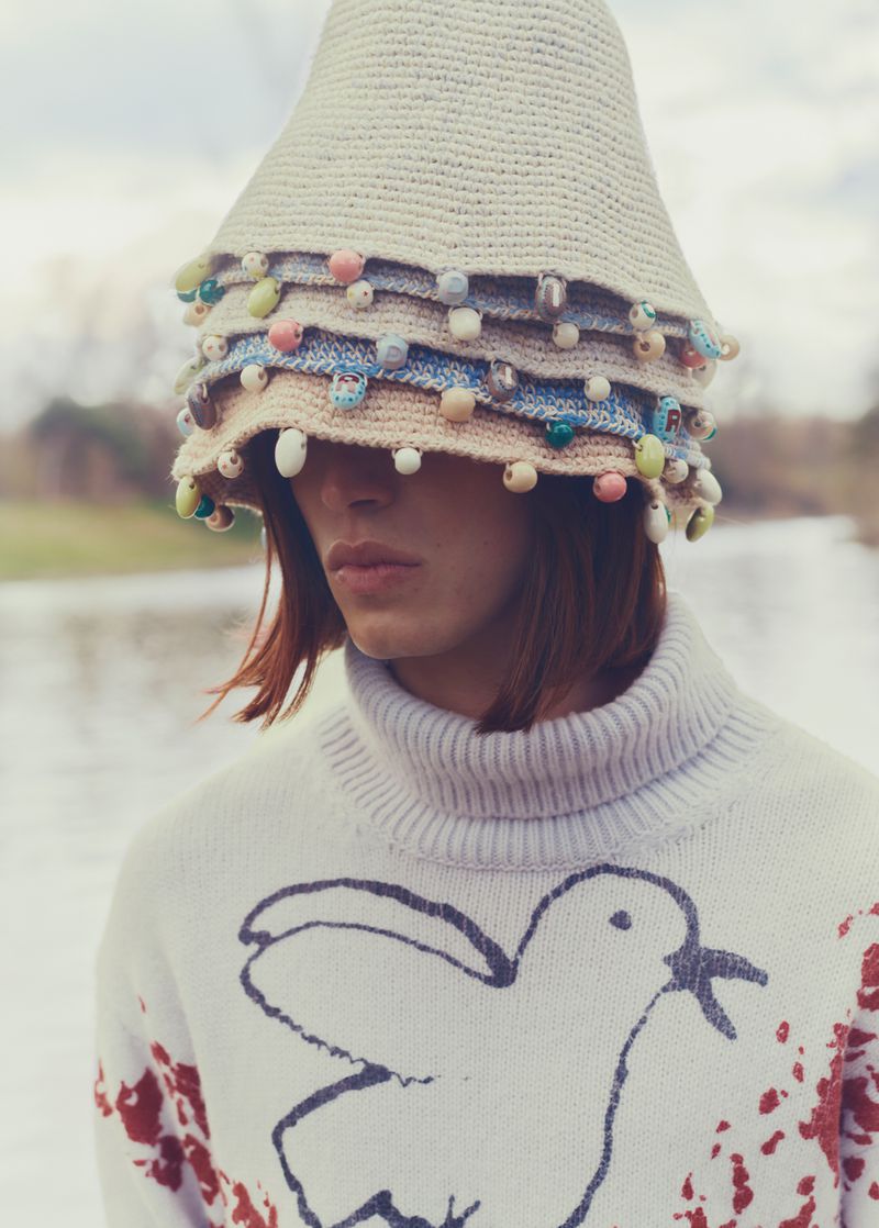 Woman in decorative knit hat and graphic sweater in outdoor setting with blurred river, trees, and cloudy sky in the background.
