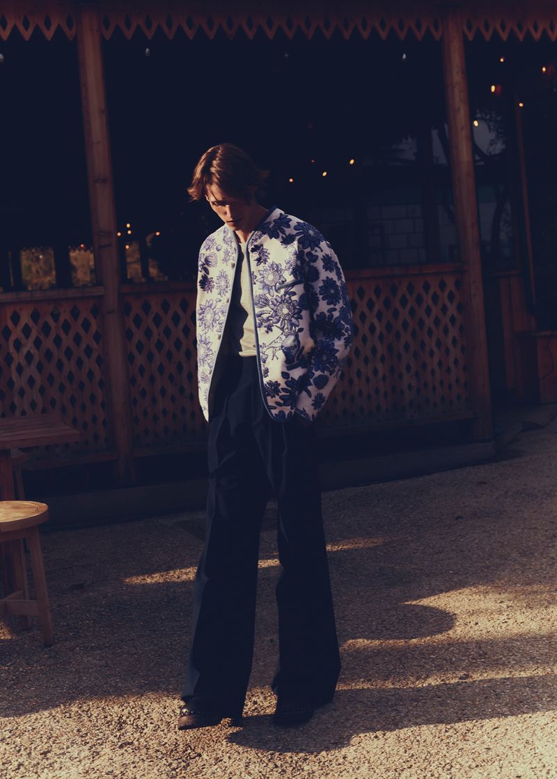 Man in floral jacket and dark pants stands on gravel in front of an open structure with hanging lights in a nighttime fashion shot.