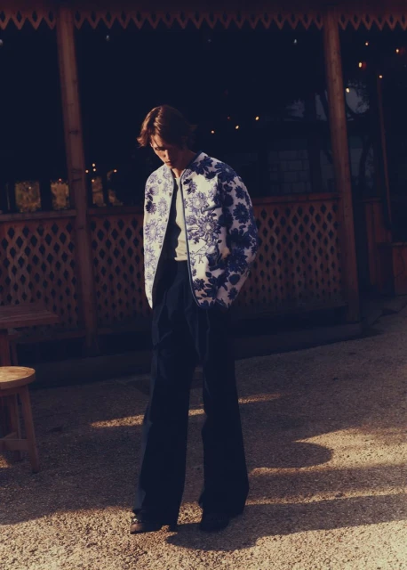 Man in floral jacket and dark pants stands on gravel in front of an open structure with hanging lights in a nighttime fashion shot.