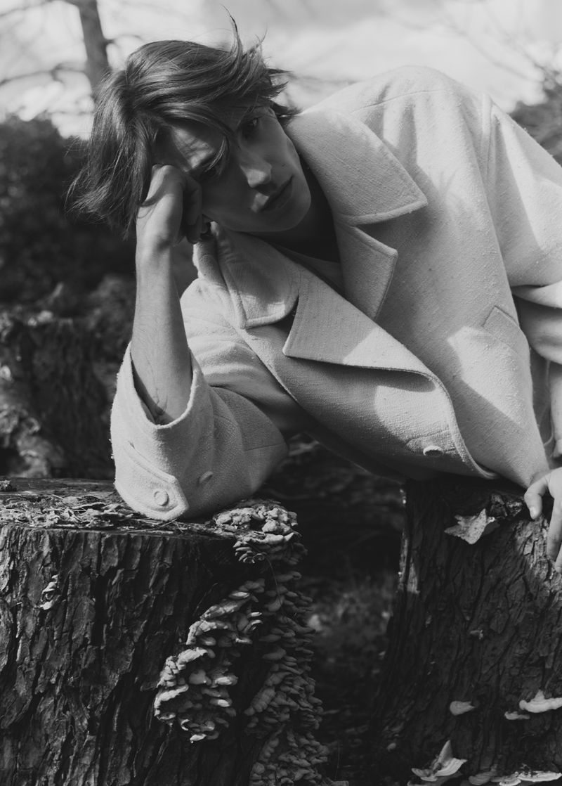 Monochrome portrait of a fair-skinned young adult male in a light-colored coat, resting his head on his hand, outdoors on a tree stump.