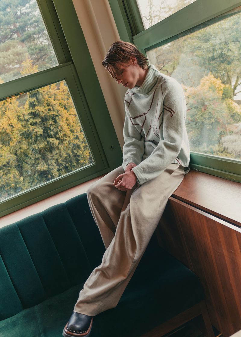 Man in oversized sweater and beige pants seated on a green couch near a window with trees in the background, fashion editorial.