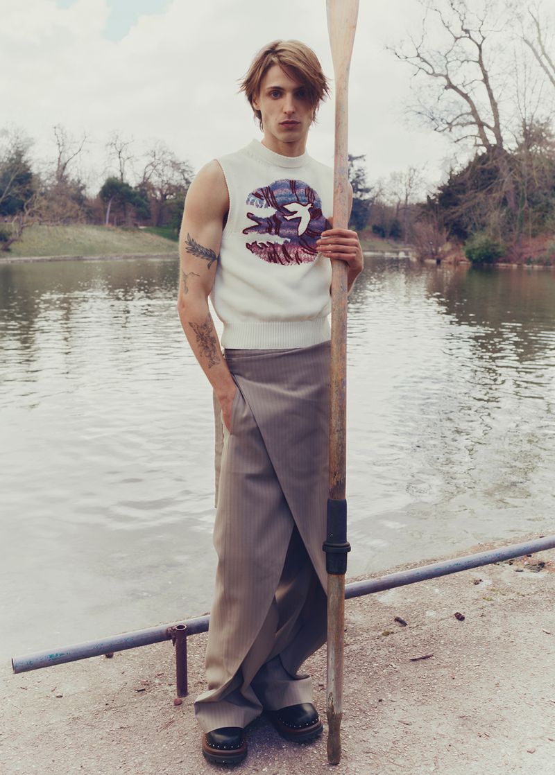 Male model stands lakeside holding oar wearing white sleeveless top and grey wrap skirt, accessorized with chunky black leather shoes.